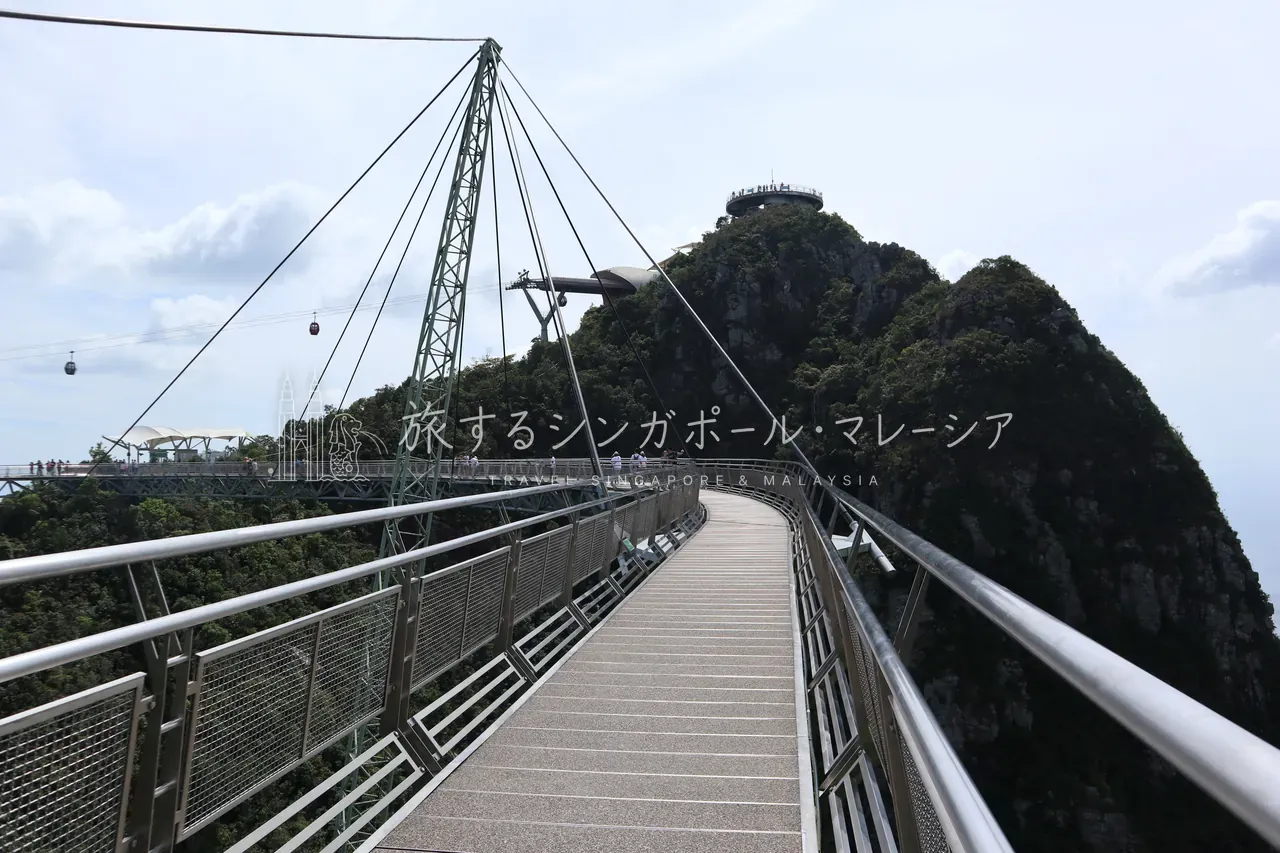 湾曲したスカイブリッジの全景。1本の支柱から扇状に張られたワイヤーで、橋が空中に弧を描きます。奥の山頂展望台、左手にはゴンドラも。これぞランカウイの象徴的な景色。