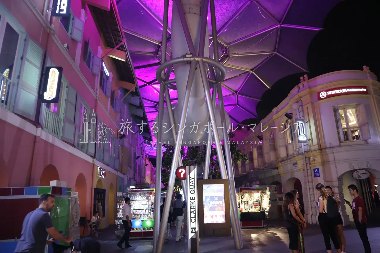 傘屋根を支える金属の支柱と、紫色に染まる屋根の裏側。足元には『CLARKE QUAY』の案内サインやエリアの店舗ガイドが立ち、パステルカラーのショップハウス（右奥は『D BLOCK』）に囲まれています。
