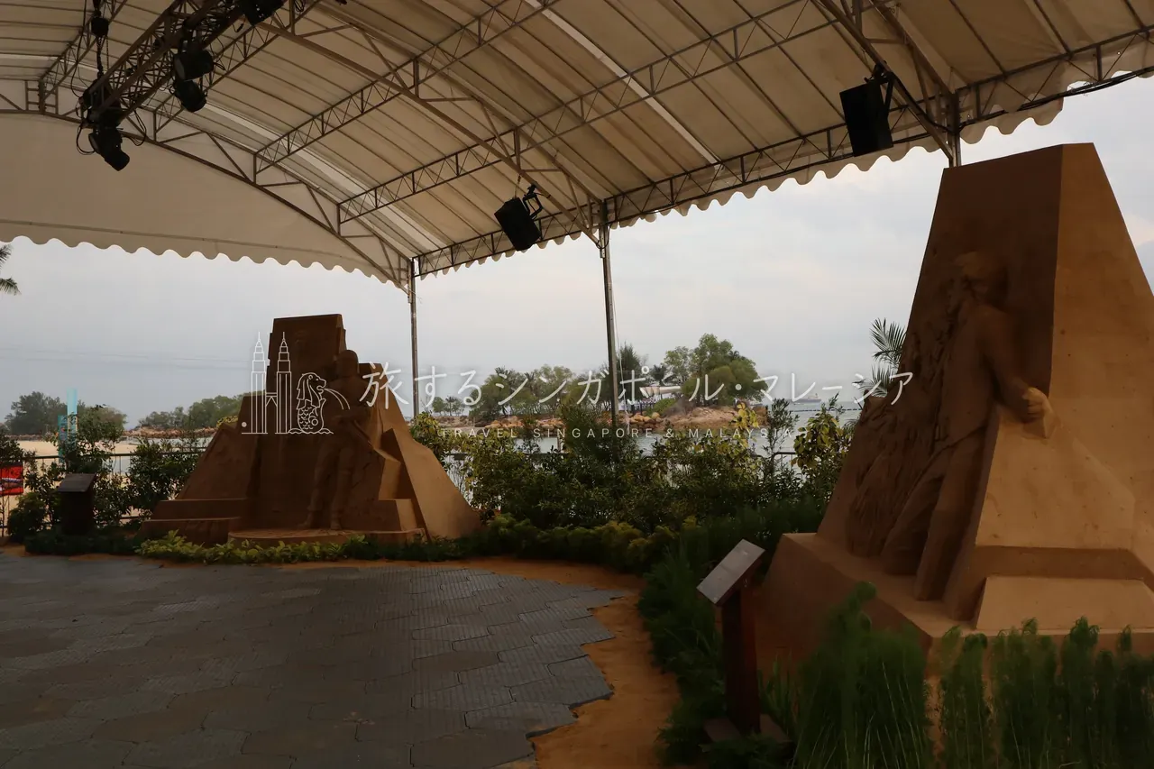 夕方の砂浜に登場していた、白いテントの下のサンドアート（砂の彫刻）。海と沖の小島を背景に、砂で作られた彫像が並びます。セントーサのビーチではこうしたイベント展示に出くわすこともあり、散歩のついでに楽しめます。