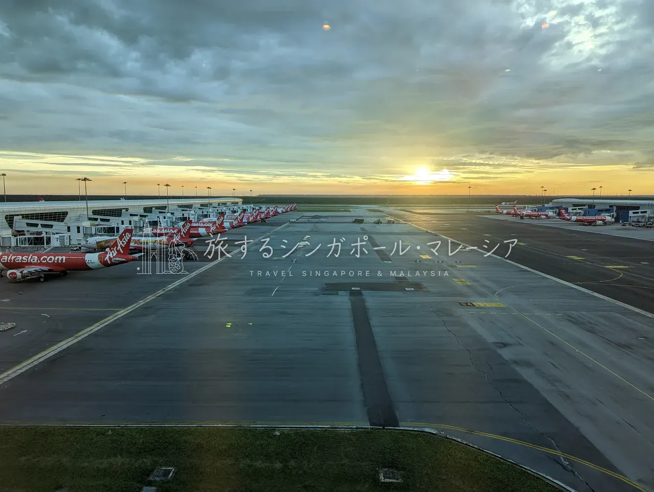 日没のklia2エプロンを、滑走路の向こうまで見渡す。左手のサテライト棟に沿って赤いAirAsia機が長い列をつくり、夕陽が誘導灯の上に沈んでいきます。LCCの一大基地が、もっとも美しく見える時間帯です。